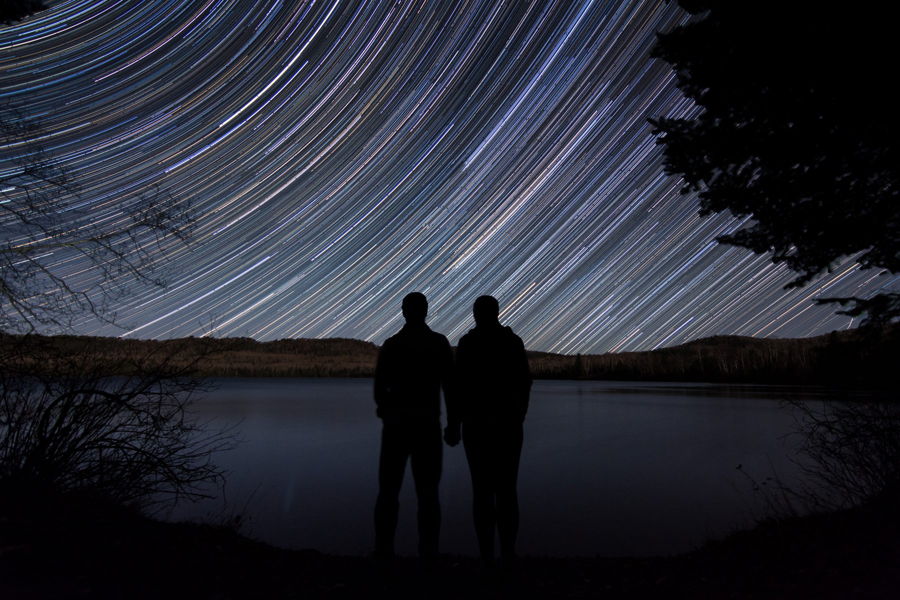 Star trails circling above two figures at the shore